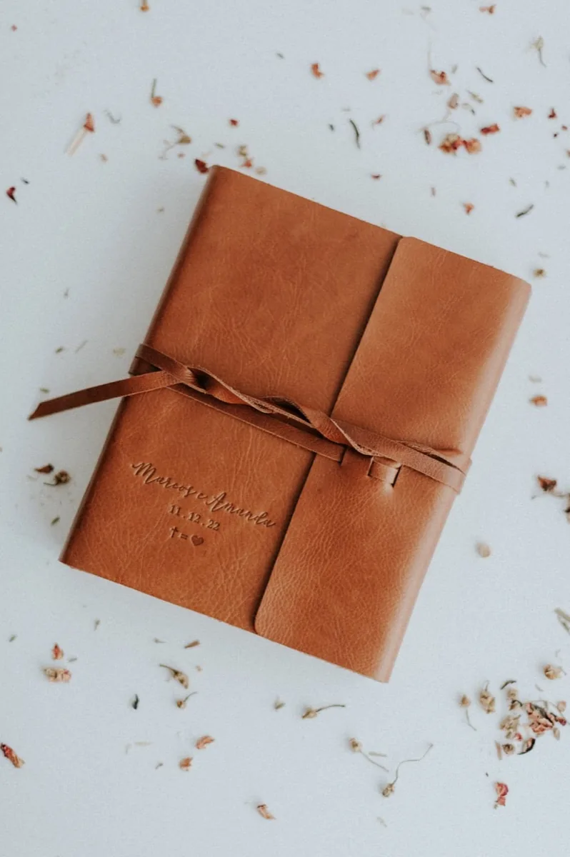 Biblia forrada en cuero café, atada con una tira de cuero y grabada con los nombres de la pareja y la fecha, sobre fondo blanco con pétalos secos