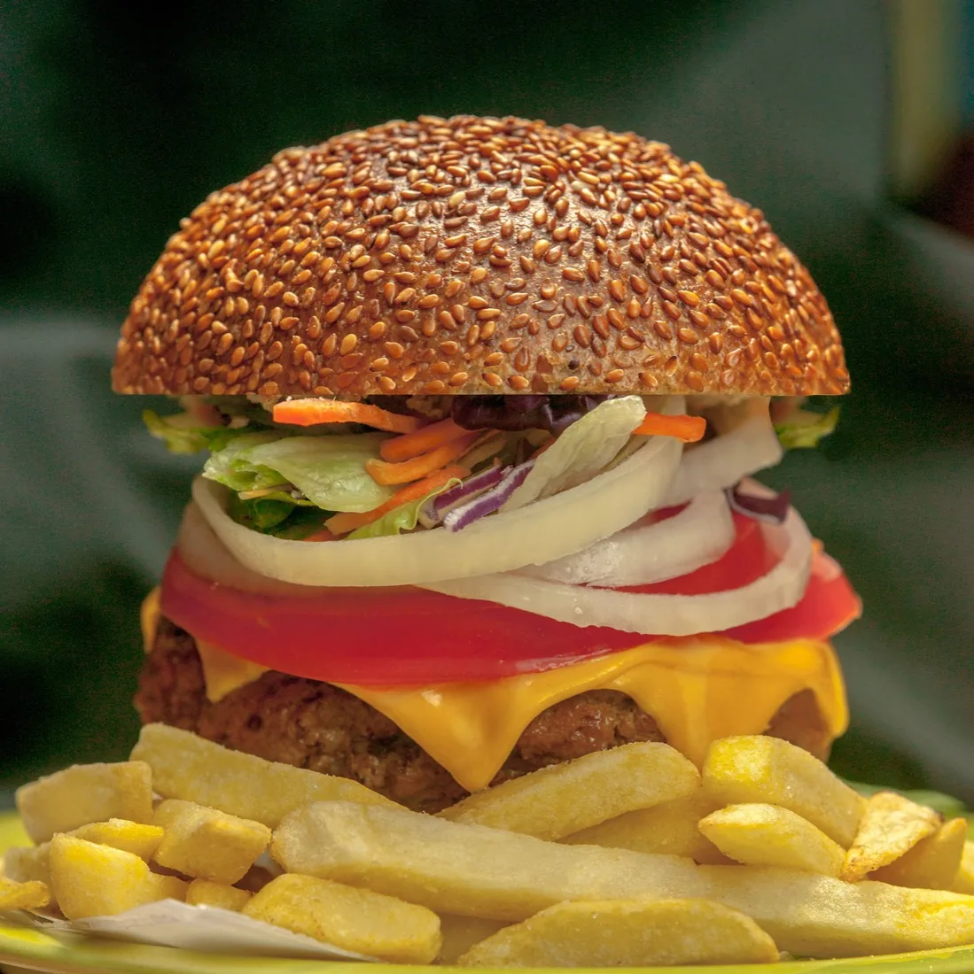 Hamburguesa con queso, tomate, cebolla y papas fritas, fotografiada con luz cálida sobre un fondo verde oscuro