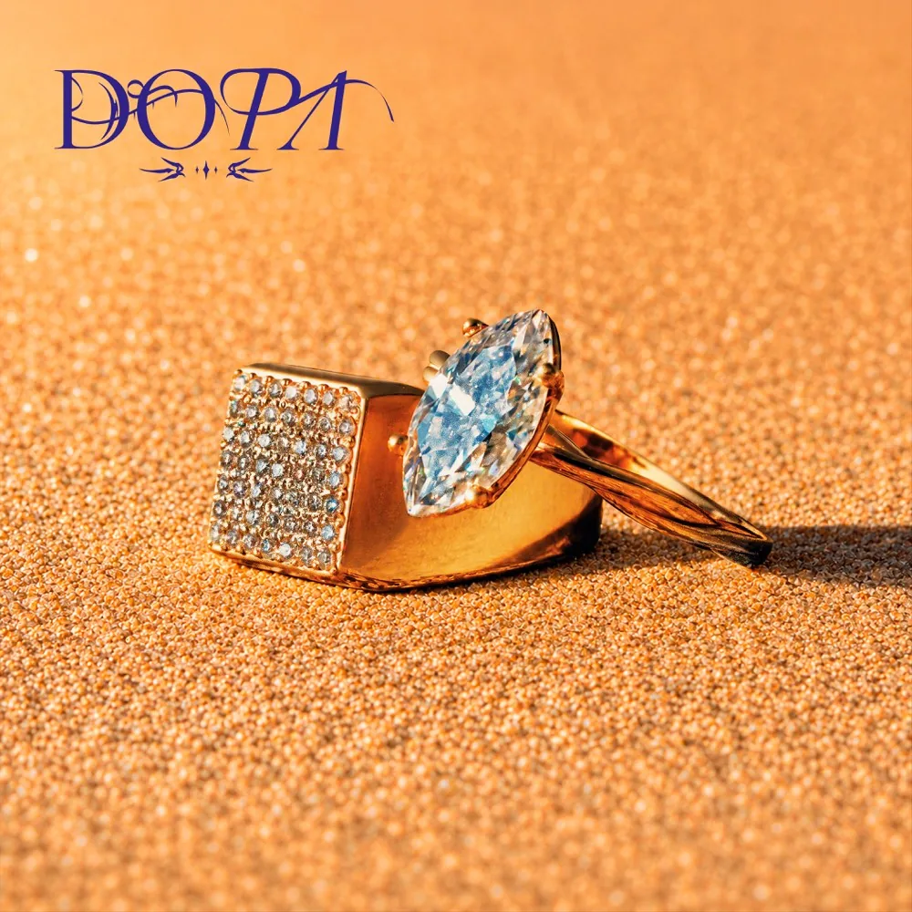 Anillo DOPA con piedra marquesa y pavé, sobre arena naranja con luz rasante y el logotipo caligráfico azul