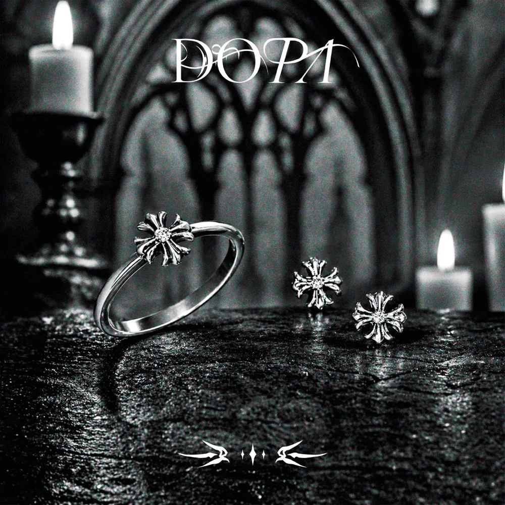 Anillo y aretes DOPA con cruz gótica sobre piedra húmeda, entre velas y un ventanal gótico, en blanco y negro con el logotipo DOPA