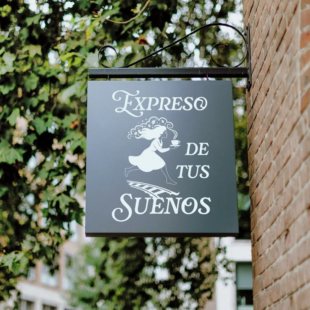 Letrero colgante de Expreso de tus Sueños en una pared de ladrillo: la soñadora corre sobre los rieles con una taza humeante