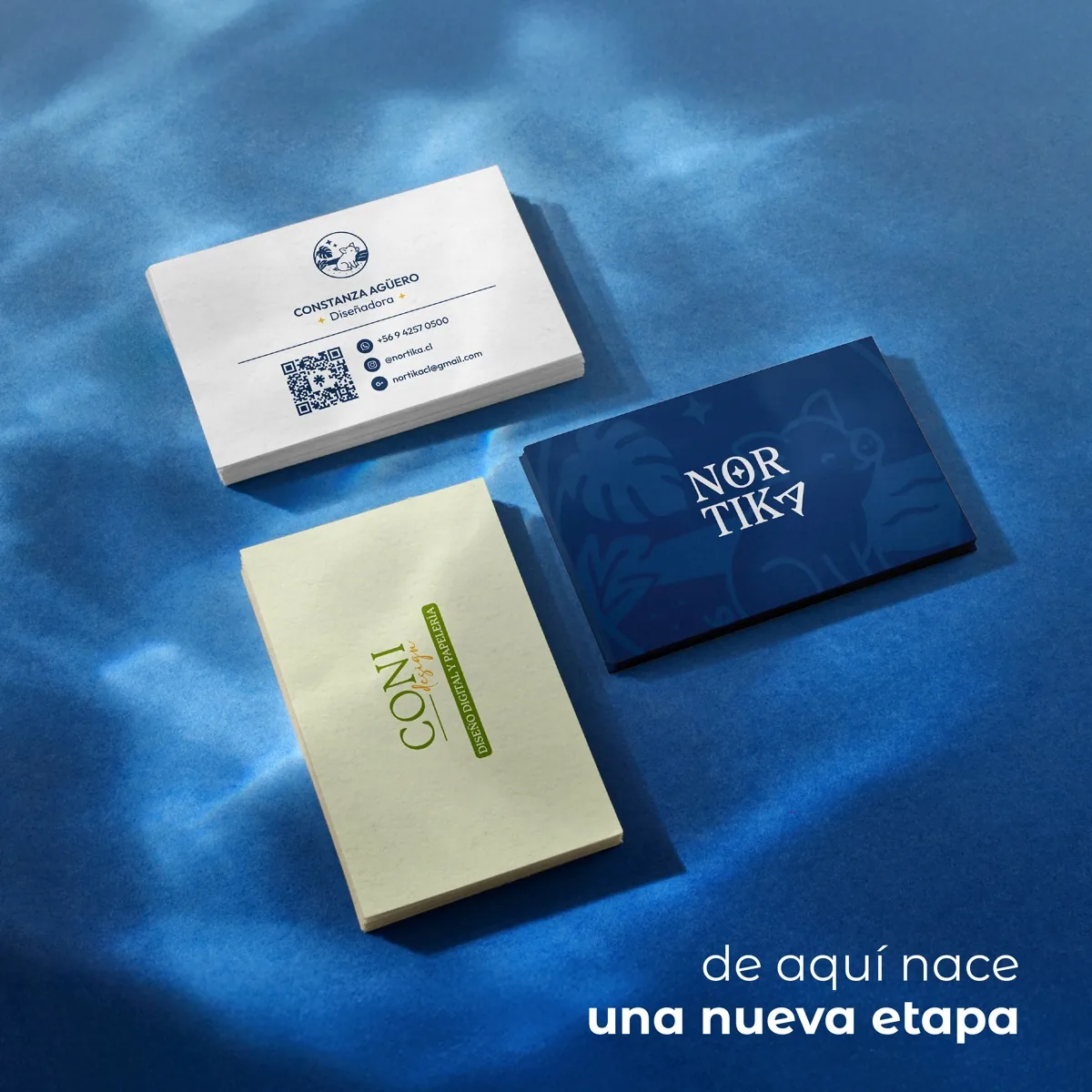 Tres pilas de tarjetas sobre papel azul con reflejos de agua: la de Coni Design en verde lima, la nueva de Nortika en azul cobalto y la de contacto de Constanza Agüero, con el texto «de aquí nace una nueva etapa»