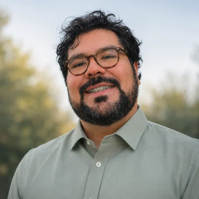 Retrato de Sebasthian Guggiana, con lentes y barba, sonriendo al aire libre con fondo verde desenfocado